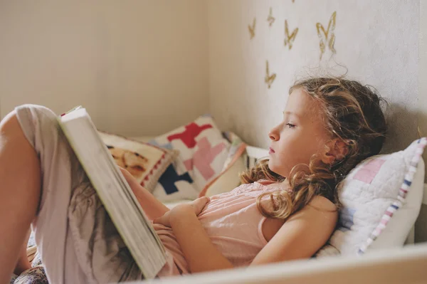 Девочка сидит на кровати и читает книгу в своей комнате. Уютные выходные дома, стиль жизни — стоковое фото