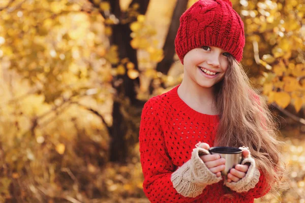 Ευτυχισμένο παιδί κορίτσι στο ζεστό κόκκινο πλεκτό καπέλο και κασκόλ για ζεστό φθινόπωρο τα πόδια πίνοντας ζεστό τσάι από θερμός. Εποχιακή υπαίθριες δραστηριότητες — Φωτογραφία Αρχείου