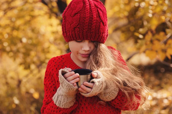 Menina feliz criança em chapéu de malha vermelho quente e cachecol na caminhada aconchegante outono beber chá quente de garrafa térmica. Atividades ao ar livre sazonais — Fotografia de Stock