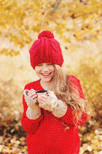 Ευτυχισμένο παιδί κορίτσι στο ζεστό κόκκινο πλεκτό καπέλο και κασκόλ για ζεστό φθινόπωρο τα πόδια πίνοντας ζεστό τσάι από θερμός. Εποχιακή υπαίθριες δραστηριότητες — Φωτογραφία Αρχείου