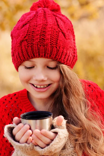 Счастливая девочка в теплой красной трикотажной шляпе и шарфе на уютной осенней прогулке пьет горячий чай из термоса. Сезонные мероприятия — стоковое фото