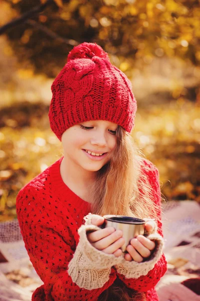 Fröhliches Kindermädchen in warmer roter Strickmütze und Schal auf einem gemütlichen Herbstspaziergang und trinkt heißen Tee aus der Thermoskanne. Saisonale Aktivitäten im Freien — Stockfoto
