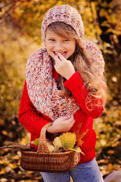 Happy child girl in knitted scarf and sweater with basket on autumn walk in forest eating apples. Fall harvest, cozy mood. — Stock Photo, Image