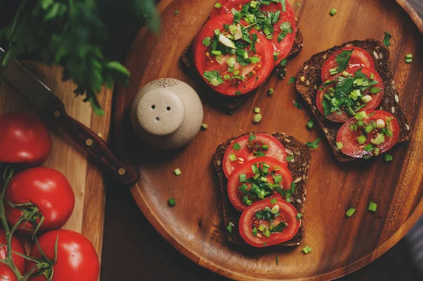 Matlagning toast smörgåsar med färsk tomat, grön lök och fullkornsbröd i mörka rustika interiören på träplatta — Stockfoto