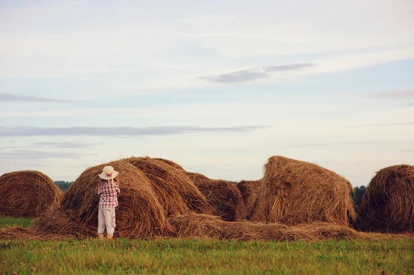 Niña feliz en estilo país camisa a cuadros y sombrero relajante en el campo de verano con pilas de heno — Foto de Stock