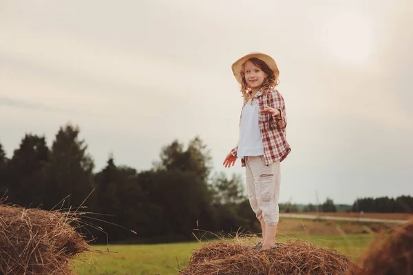 Niña feliz en estilo país camisa a cuadros y sombrero relajante en el campo de verano con pilas de heno — Foto de Stock