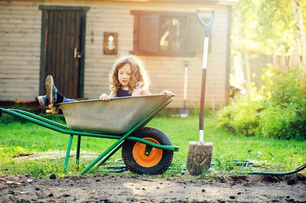 Happy kid girl playing in summer garden with wheelbarrow and shovel and helping — Stock Photo, Image