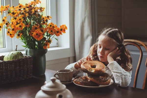 Bambina che fa colazione a casa in autunno mattina. Vita reale accogliente interni moderni in casa di campagna. Bambino mangiare bagel e bere tè . — Foto Stock