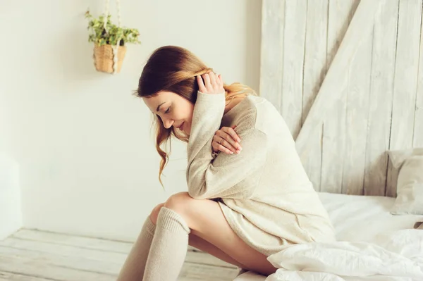 Νεαρή γυναίκα στην πλεκτή ζακέτα και ζεστά κάλτσες wake μέχρι το πρωί στο άνετο υπνοδωμάτιο Σκανδιναβική και κάθονται στα κρεβάτια με λευκά κλινοσκεπάσματα. Casual τρόπου ζωής σε μοντέρνο εσωτερικό — Φωτογραφία Αρχείου