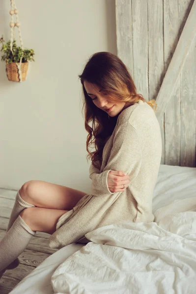 Young woman in knitted cardigan and warm socks wake up in the morning in cozy scandinavian bedroom and sitting on bed with white bedlinen. Casual lifestyle in modern interior — Stock Photo, Image