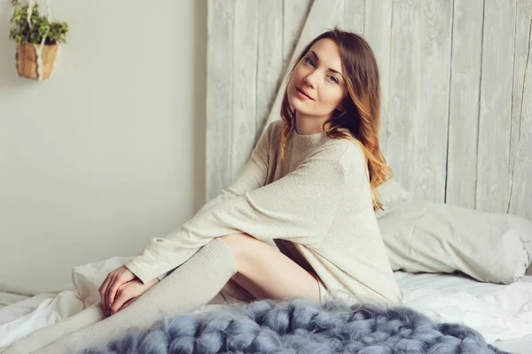 Young woman in knitted cardigan and warm socks wake up in the morning in cozy scandinavian bedroom and sitting on bed with white bedlinen. Casual lifestyle in modern interior — Stock Photo, Image