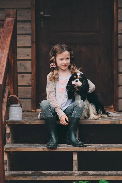 Kid girl playing with her spaniel dog, sitting on stairs at wooden log cabin — Stock Photo, Image