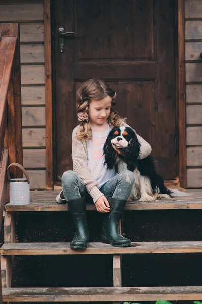 Kid girl playing with her spaniel dog, sitting on stairs at wooden log cabin — Stock Photo, Image