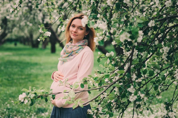 Gelukkig zwangere blonde mooie vrouw lopen ondersteunt in voorjaar park of tuin, poseren in de buurt van bloeiende boom — Stockfoto