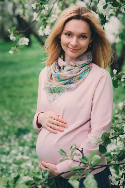 Happy pregnant blonde beautiful woman walking outddor in spring park or garden, posing near blooming tree — Stock Photo, Image