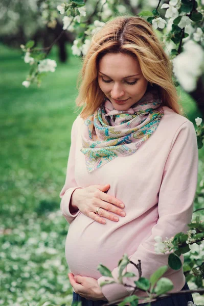 Happy pregnant blonde beautiful woman walking outddor in spring park or garden, posing near blooming tree — Stock Photo, Image