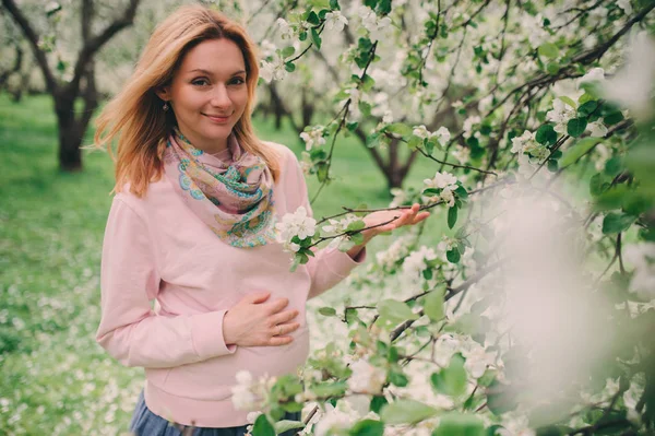 Szczęśliwy w ciąży Blondynka piękna kobieta spaceru outddor w parku lub ogrodzie, pozowanie w pobliżu kwitnące drzewo — Zdjęcie stockowe