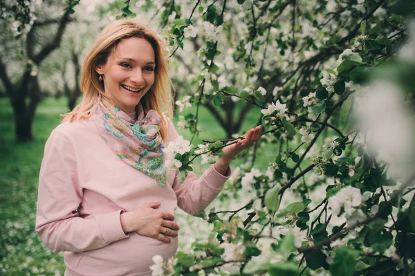 Happy pregnant blonde beautiful woman walking outddor in spring park or garden, posing near blooming tree — Stock Photo, Image