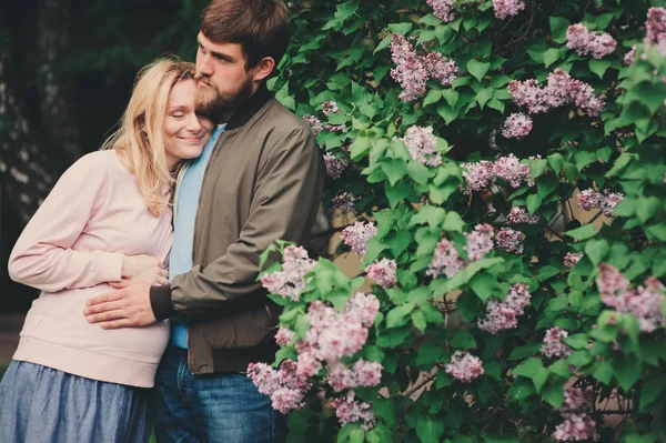 Glückliches Liebespaar mit schwangerer Frau, die im Frühlingspark spaziert und in der Nähe blühender Lilienbüsche posiert — Stockfoto