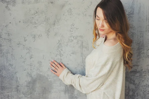 Νεαρή γυναίκα στο υπερμεγέθης πλεκτά πουλόβερ που παρουσιάζουν από τον γκρίζο τοίχο σε μοντέρνο διαμέρισμα. Casual τρόπου ζωής στο σπίτι — Φωτογραφία Αρχείου