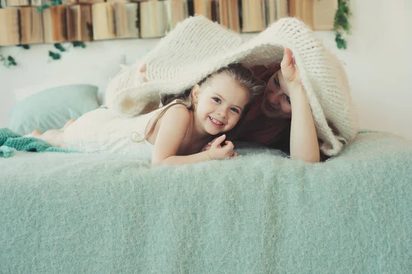 Счастливая семья играет дома. Мать и малышка дочь расслабляются и веселятся в постели в уютные выходные утром — стоковое фото