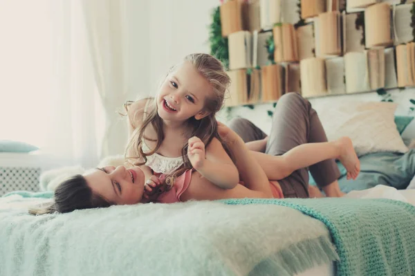 Счастливая семья играет дома. Мать и малышка дочь расслабляются и веселятся в постели в уютные выходные утром — стоковое фото