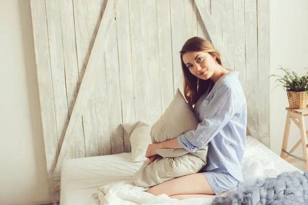 Mujer joven en pijama se despierta por la mañana en el acogedor dormitorio escandinavo y se sienta en la cama con ropa de cama blanca. Estilo de vida casual en el interior moderno — Foto de Stock