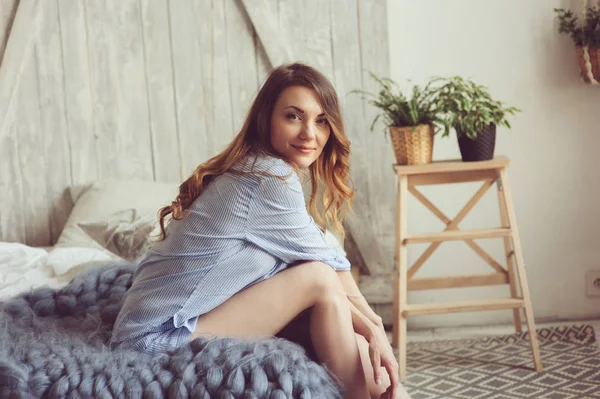 Mujer joven en pijama se despierta por la mañana en el acogedor dormitorio escandinavo y se sienta en la cama con plantas de interior en el fondo. Estilo de vida casual en el interior moderno — Foto de Stock