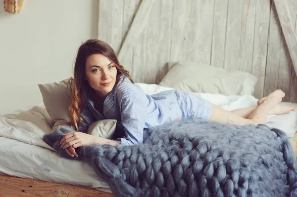 在舒适的斯堪的纳维亚卧室和坐在床上用白色床单睡衣的年轻女人在早上醒来。在现代室内的休闲生活方式 — 图库照片
