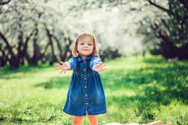 Spring portrait of cute little toddler girl in blue jeans dress walking in blooming park or garden — Stock Photo, Image