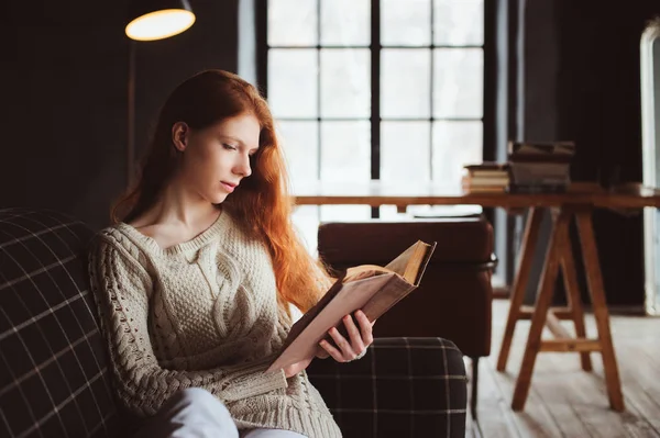 Joven hermosa pelirroja mujer relajarse en casa en el otoño acogedora noche y leer libro en el sofá —  Fotos de Stock