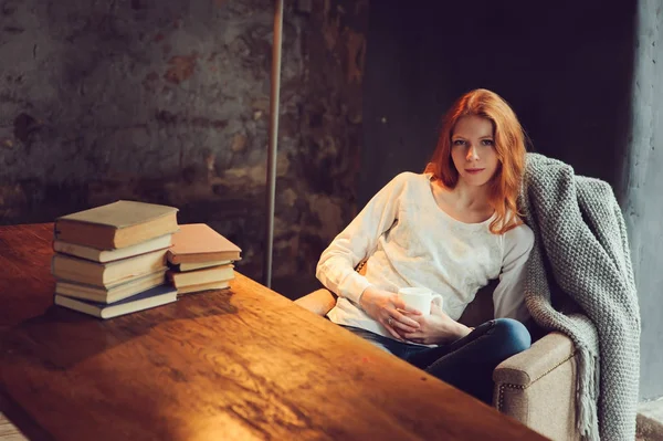 Joven hermosa pelirroja que se relaja en la silla en casa con taza de té o cacao. Acogedor concepto de otoño por la noche . — Foto de Stock