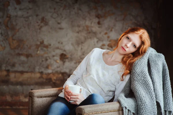 Wanita muda berambut merah yang cantik santai di kursi di rumah dengan secangkir teh atau coklat. Konsep malam musim gugur yang nyaman . — Stok Foto