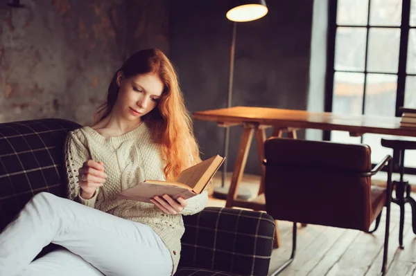 Joven hermosa pelirroja mujer relajarse en casa en el otoño acogedora noche y leer libro en el sofá — Foto de Stock