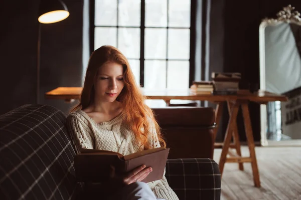 Jeune belle rousse femme se détendre à la maison dans la soirée d'automne confortable et livre de lecture sur le canapé — Photo