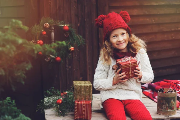 Bambina felice con cappello rosso e sciarpa che avvolge i regali di Natale in un'accogliente casa di campagna, decorata per Capodanno e Natale. Preparativi per vacanze con bambini . — Foto Stock