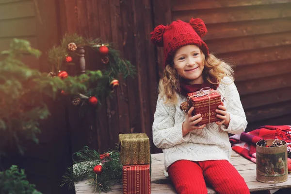 Bambina felice con cappello rosso e sciarpa che avvolge i regali di Natale in un'accogliente casa di campagna, decorata per Capodanno e Natale. Preparativi per vacanze con bambini . — Foto Stock