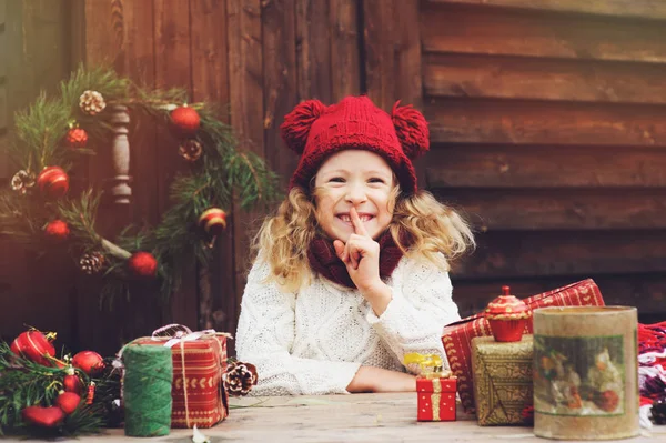 Happy child girl in red hat and scarf wrapping Christmas gifts at cozy country house, decorated for New Year and Christmas. Preparations for holidays with kids. — Stock Photo, Image