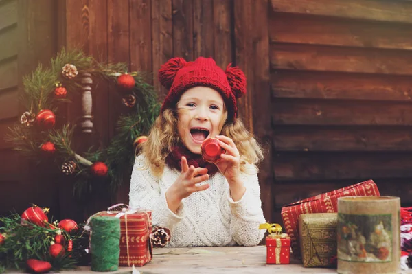 Bambina felice con cappello rosso e sciarpa che avvolge i regali di Natale in un'accogliente casa di campagna, decorata per Capodanno e Natale. Preparativi per vacanze con bambini . — Foto Stock