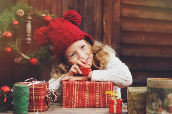 Bambina felice con cappello rosso e sciarpa che avvolge i regali di Natale in un'accogliente casa di campagna, decorata per Capodanno e Natale. Preparativi per vacanze con bambini . — Foto Stock