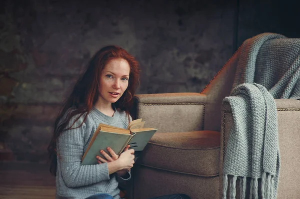 Heureuse rousse femme détente à la maison en hiver confortable ou week-end d'automne avec livre et tasse de thé chaud, assis dans une chaise douce avec couverture — Photo