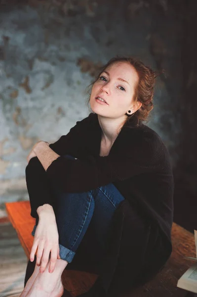 Young hipster redhead woman relaxing at home, sitting on wooden table with books in modern loft interior — Stock Photo, Image