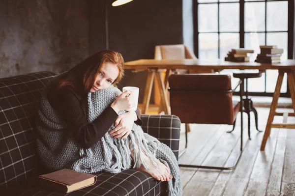 Wanita muda sakit penyembuhan dengan minuman panas di rumah di sofa nyaman, dibungkus dengan selimut rajutan — Stok Foto