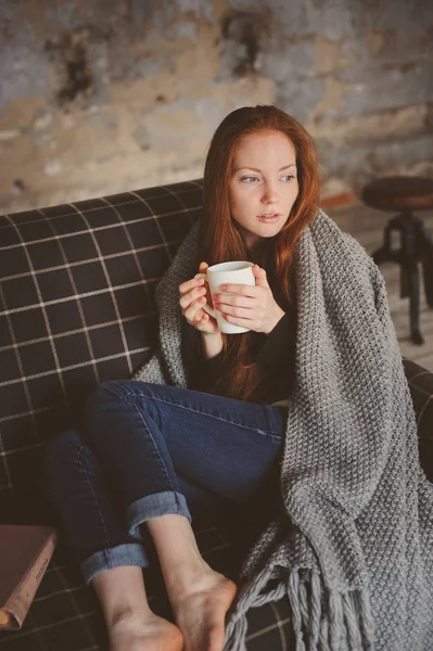 Wanita muda sakit penyembuhan dengan minuman panas di rumah di sofa nyaman, dibungkus dengan selimut rajutan — Stok Foto