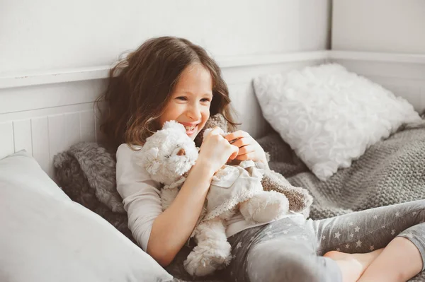 Szczęśliwy dziewczyna bawi się misie w jej pokoju, siedząc na łóżku w piżamy — Zdjęcie stockowe