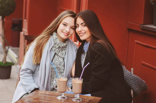 Twee gelukkige meisje vrienden praten en drinken koffie in herfst stad in café. Vergadering van goede vrienden, jonge modieuze studenten met natuurlijke make-up. — Stockfoto