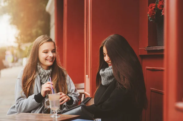 Twee gelukkige meisje vrienden praten en drinken koffie in herfst stad in café. Vergadering van goede vrienden, jonge modieuze studenten met natuurlijke make-up. — Stockfoto