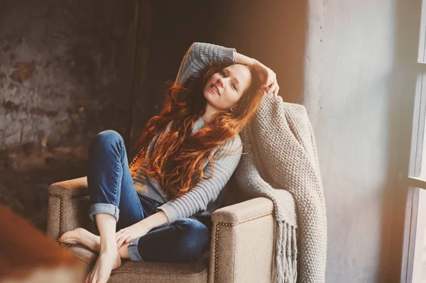 Joven readhead mujer relajante en casa en silla acogedora, vestido con suéter casual y jeans. Calma fin de semana de invierno u otoño —  Fotos de Stock