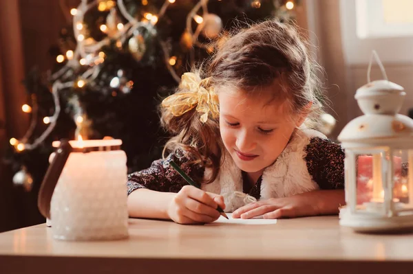 Kız Çocuk evde Noel Baba'ya mektup yazma. 8 yıl yaşlı kız Noel ve yeni yıl evde ağaç arka plan üzerinde süslenmiş için hediye listesi yapma