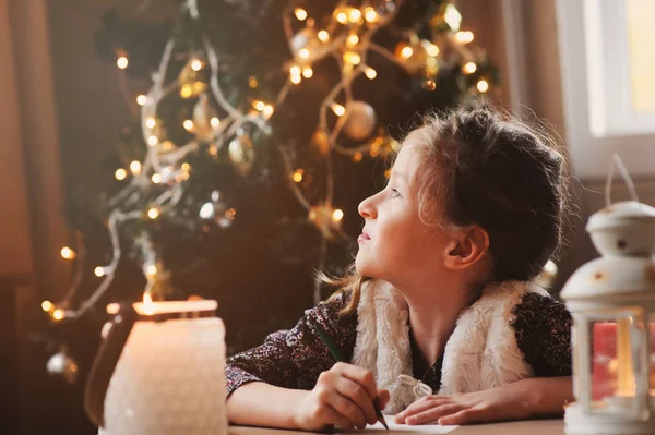 Kız Çocuk evde Noel Baba'ya mektup yazma. 8 yıl yaşlı kız Noel ve yeni yıl evde ağaç arka plan üzerinde süslenmiş için hediye listesi yapma